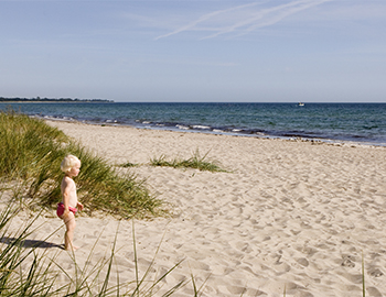 Lille pige står ved klitterne på sandstrand og kigger ud over havet.